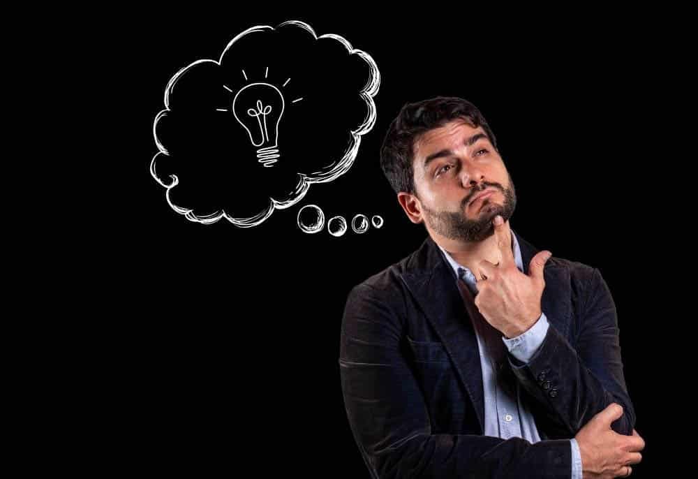 Homem pensativo com fundo preto, apontando para o rosto, enquanto uma ilustração de um balão de pensamento com uma lâmpada aparece ao lado de sua cabeça, simbolizando uma ideia criativa. Representação conceitual de novas ideias para renda extra e oportunidades financeiras.